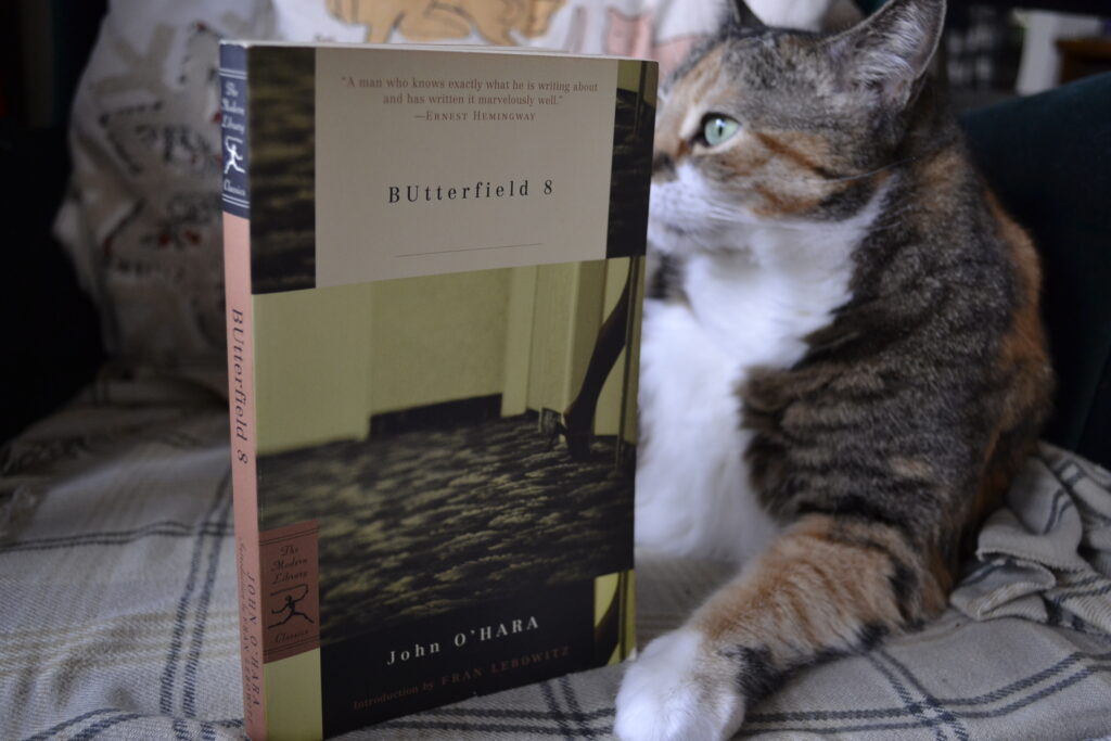 A calico cat sits behind a copy of BUtterfield 8.