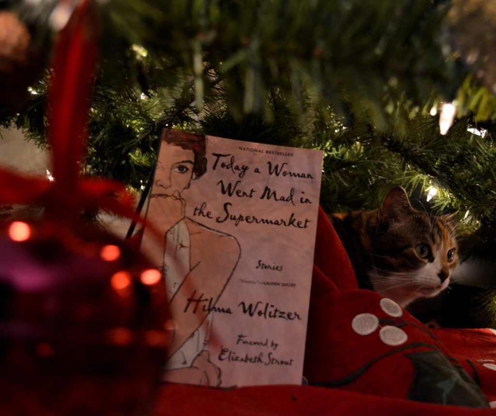 A copy of Today a Woman Went Mad in the Supermarket sits beneath a Christmas tree with a wide-eyed tabby cat.