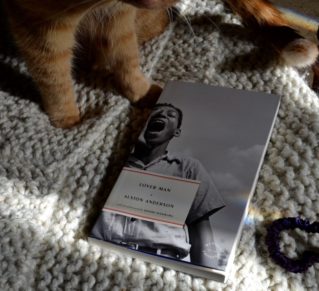 A black and white book sits on a knitted blanket. Just the paws of an orange cat are visible beside it.