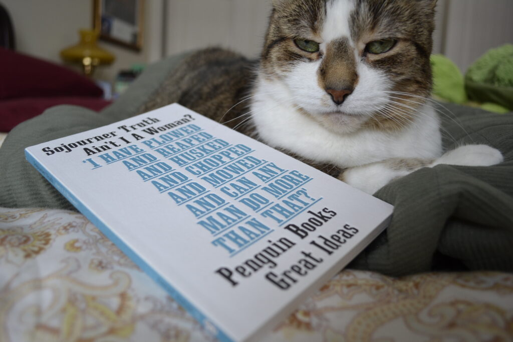 A cat looks sleepily at the camera beside a copy of Sojourner Truth's Ain't I a Woman?