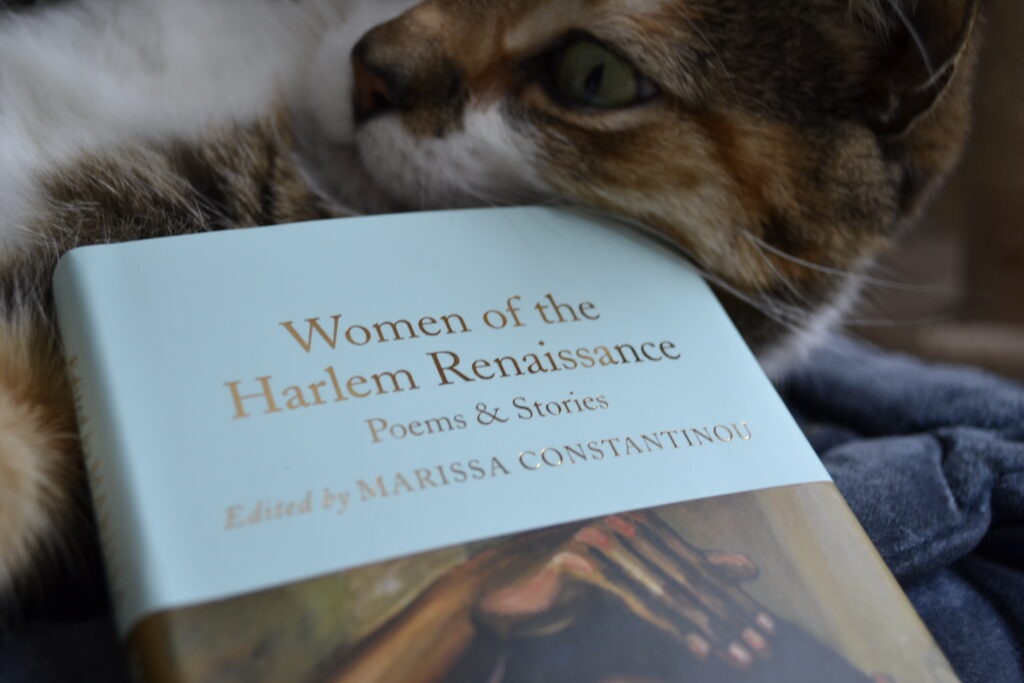 A calico tabby rubs the top of a blue book: Women of the Harlem Renaissance, Poems & Stories, edited by Marissa Constantinou.