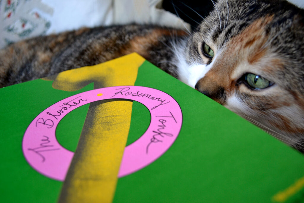 A calico tabby sniffs a book. A yellow finger on the book pokes through a pink ring that reads: The Bloater * Rosemary Tonks.