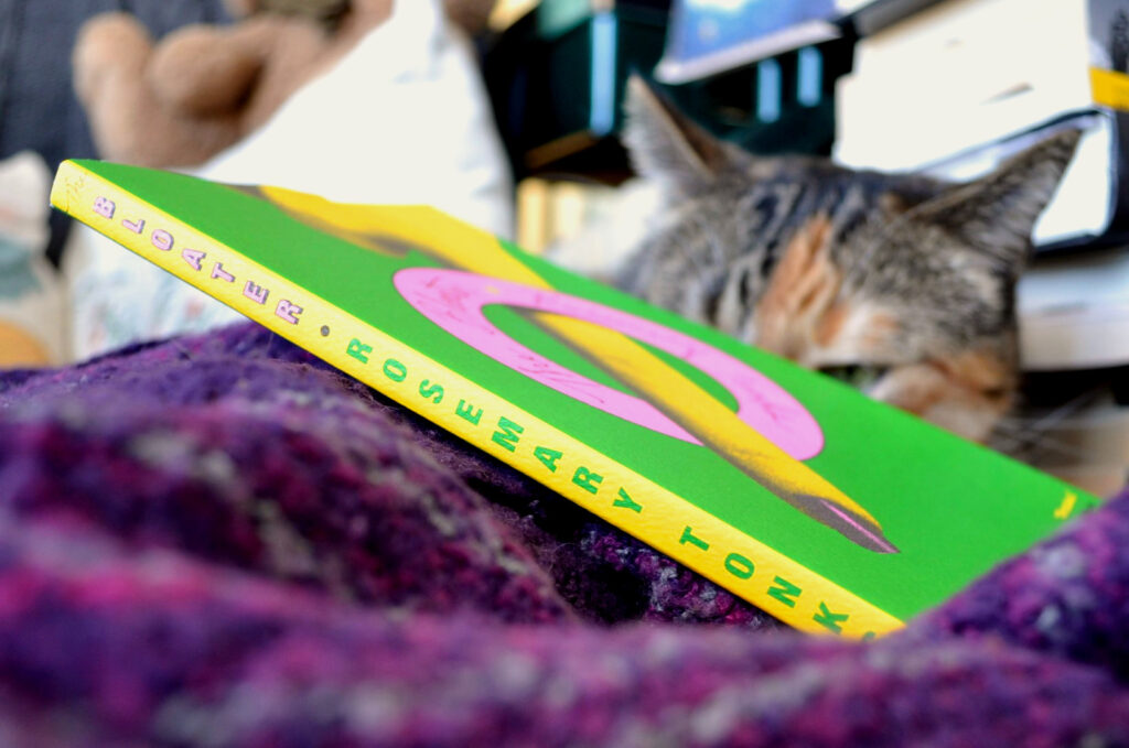 Behind the vibrantly yellow spine of The Bloater, a calico tabby cat can be seen.
