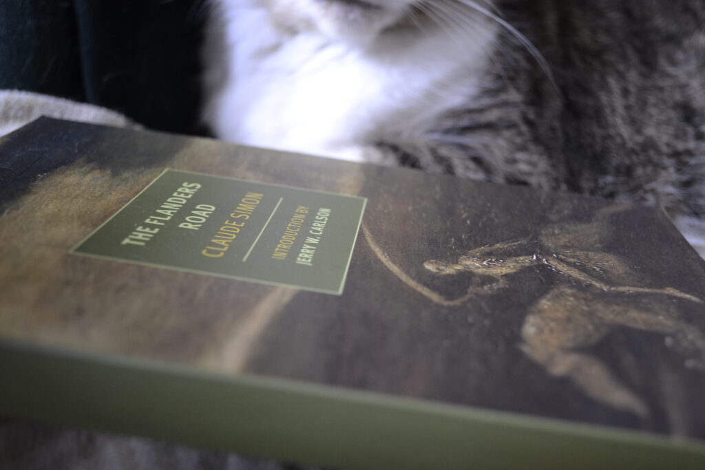 The Flanders Road by Claude Simon rests on its side. The green spine and the cover can be seen. The cover shows a skeleton with a scythe riding a horse.