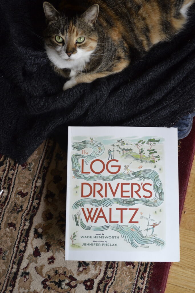 A calico cat lies beside a copy of The Log Driver's Waltz. The cover is illustrated with a log driver bringing logs down the river.