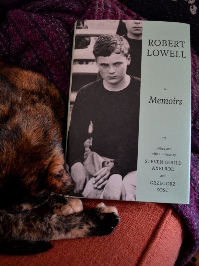 A tortoiseshell cat curls around a copy of Robert Lowell's Memoirs.