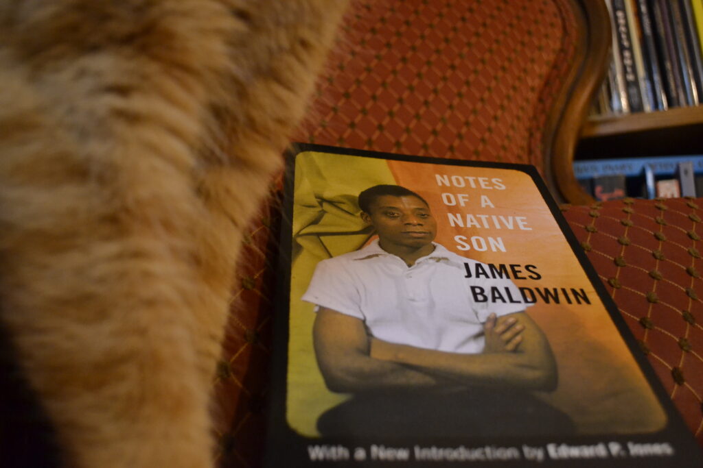 Baldwin's Notes of a Native Son has a cover with an image of Jame Baldwin on the cover, his arms crossed, wearing a white shirt and black pants.