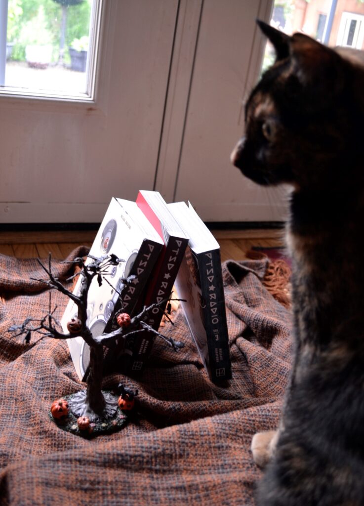 A tortie looks perturbed beside three stacked volumes of PTSD Radio.