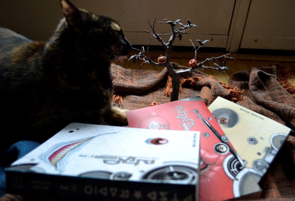 A tortie sniffs a miniature tree covered with jack-o-lanterns and bars. Beside her lay three creepy books.