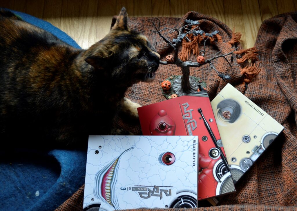 A tortie chews a little halloween tree beside three volumes of PTSD Radio.