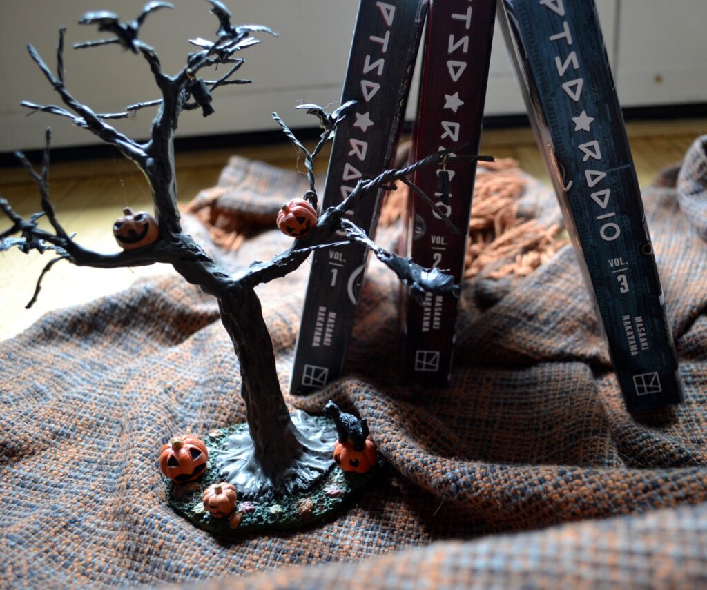 Three volumes of PTSD Radio lean together above a miniature tree decorated with jack-o-lanterns and bats.