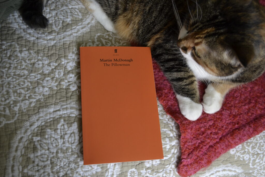 A calico tabby with white paws curls around the edge of an orange book, The Pillowman.