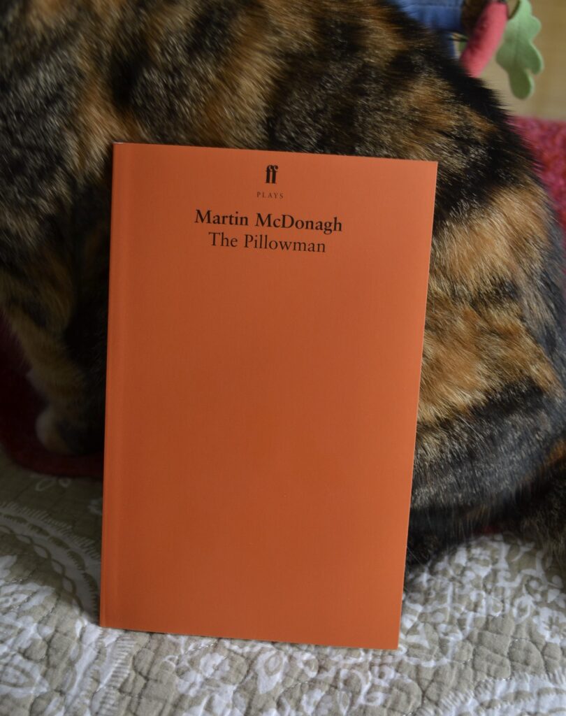 The Pillowman by Martin McDonagh is a plain orange book. It lies against the side of a tabby cat.