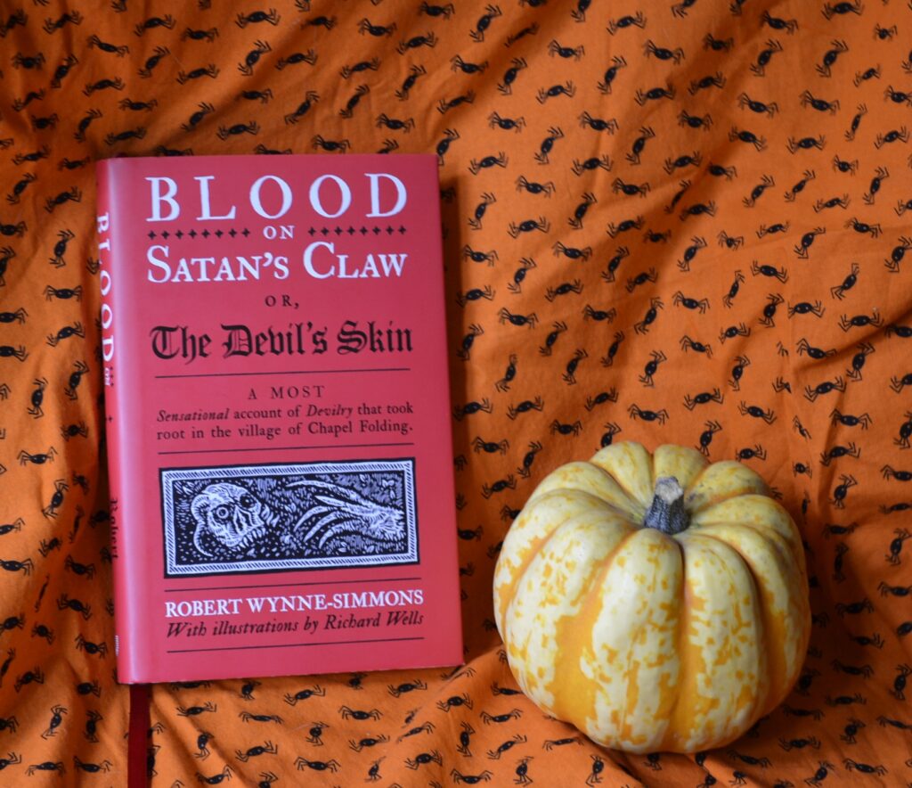 A red book — Blood on Satan's Claw — sits on an orange spider-patterned background with a small yellow squash with orange stripes.