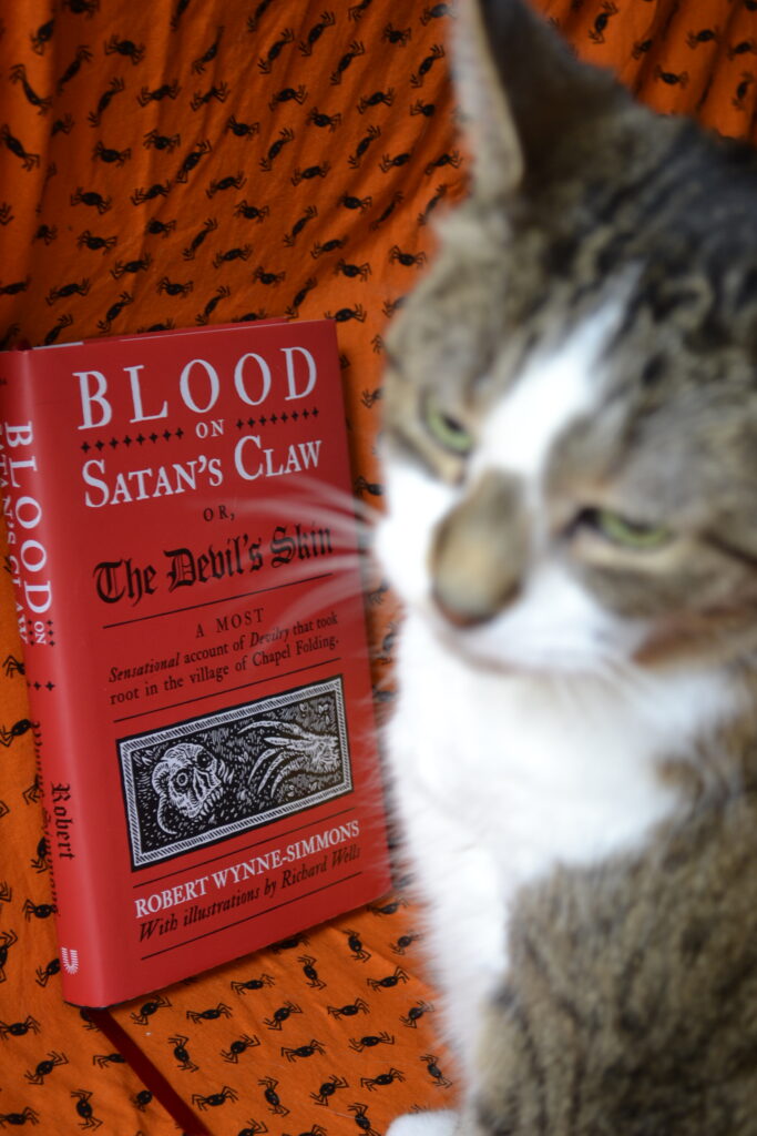 A tabby cat with a brown nose and big ears site beside a red book: Blood on Satan's Claw.