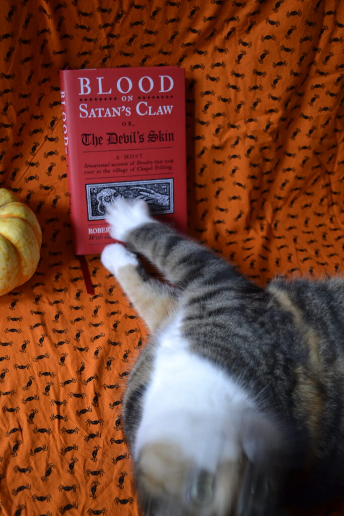 A cat pushes away a red book. The text on the book's cover reads: Blood on Satan's Claw or The Devil's Skin — a most sensational account of Devilry that took root in the village of Chapel Folding.