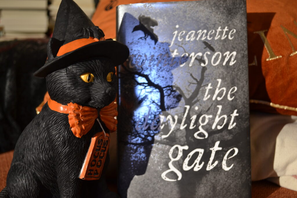 A statue of a black cat in a witch hat sits beside a grey book, The Daylight Gate.