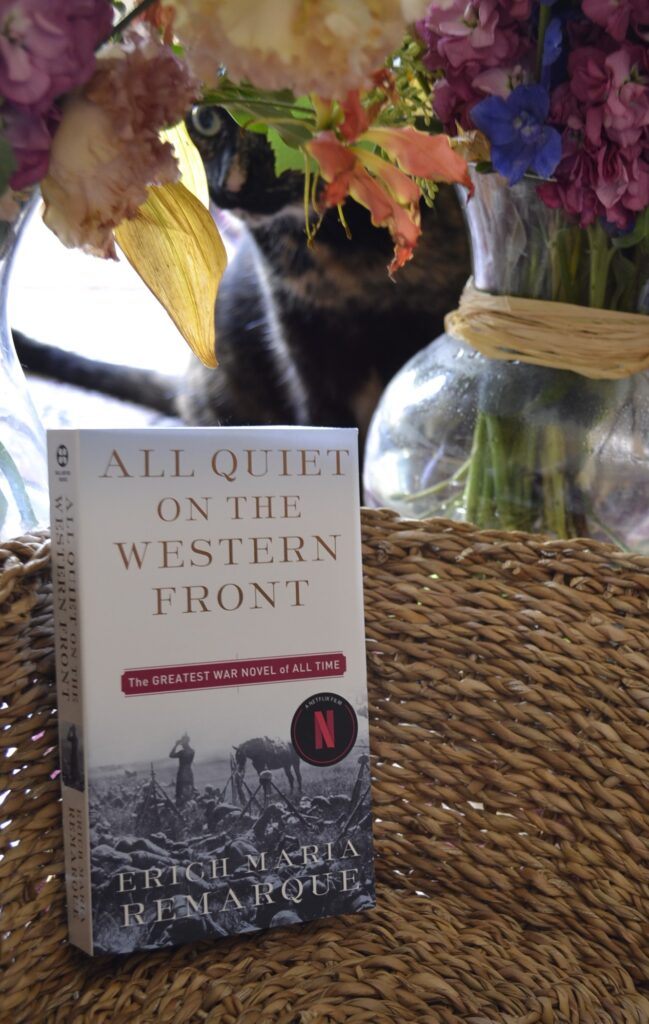 A tortie peeks out from beneath some flowers. In front of her is a book: All Quiet on the Western Front.