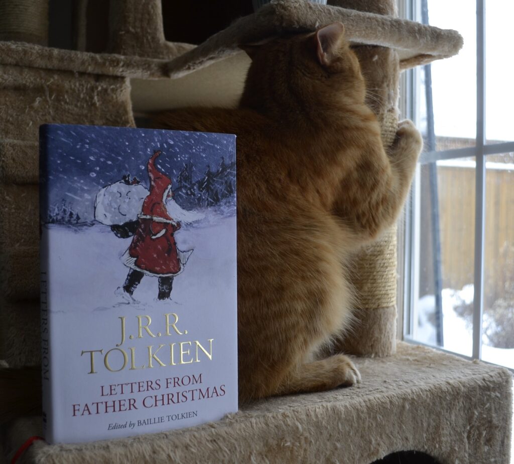 An orange tabby cat climbs a cat tree. Beside her is a book that has a hand-drawn image of Father Christmas on it.