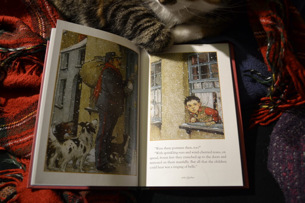 An open book shows two pictures: one of a postman with a red scarf knocking on a door with two dogs and a cat beside him; the other of a young boy leaning out an open window.