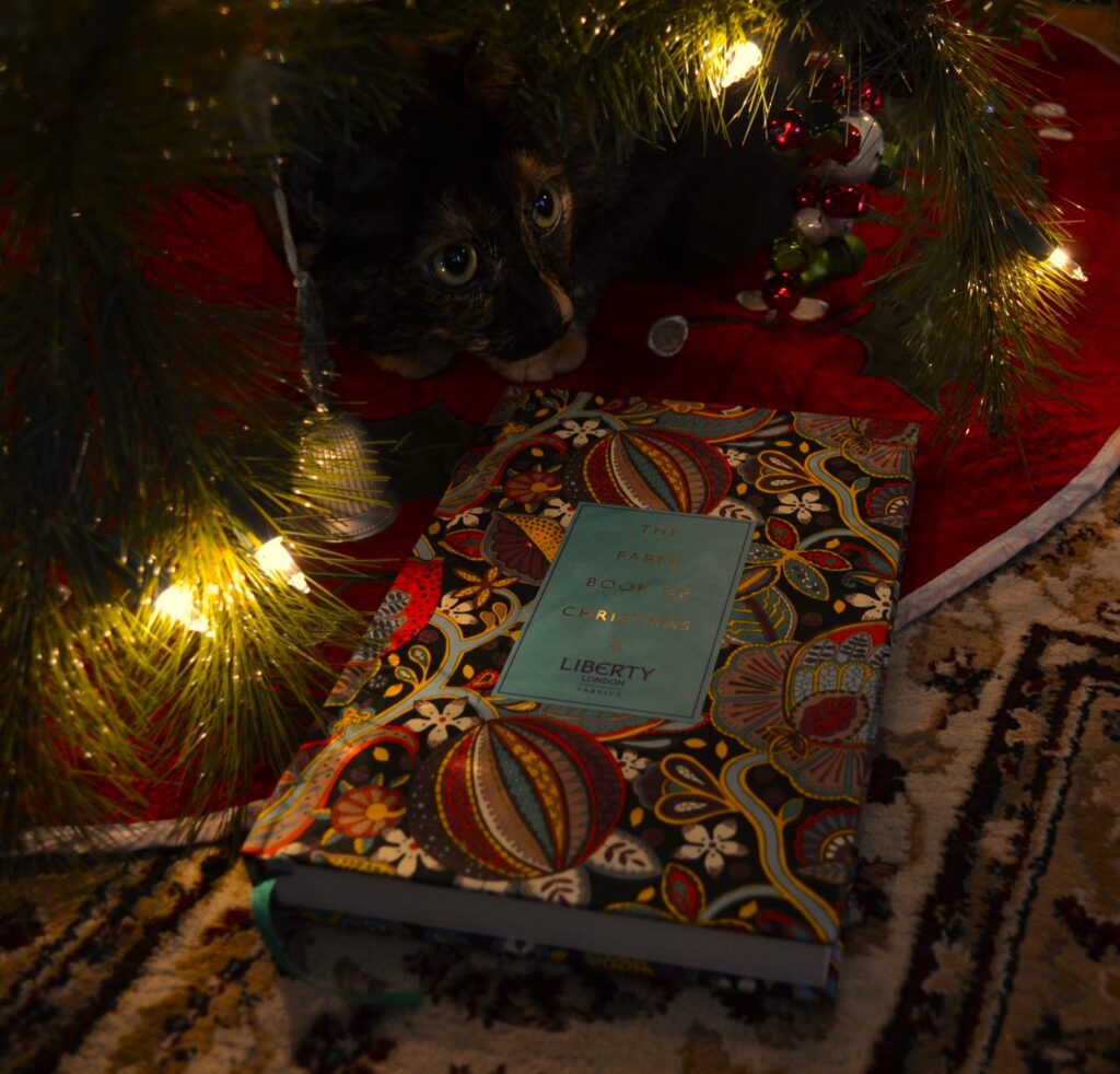 A tortoiseshell cat sits sweetly beneath the glow of a Christmas tree. In front of her is The Faber Book of Christmas.