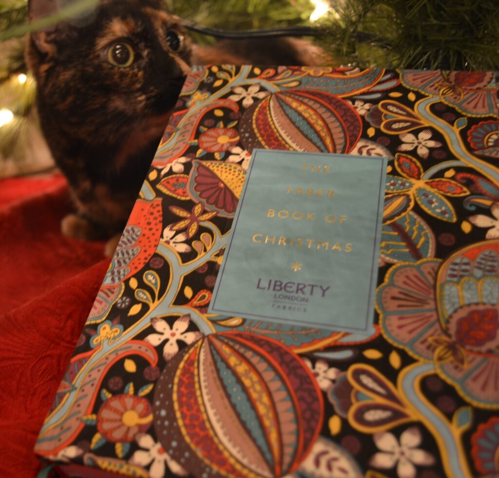 The Faber Book of Christmas sits beneath a Christmas tree, in the glow of the lights. A tortie lurks behind it.