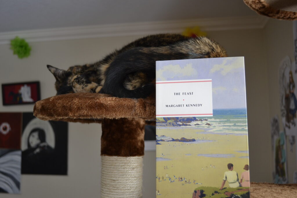 A tortoiseshell cat sleep on a cat tree. Beside her is The Feat by Margaret Kennedy. The Feast has a muted beach scene on the cover.