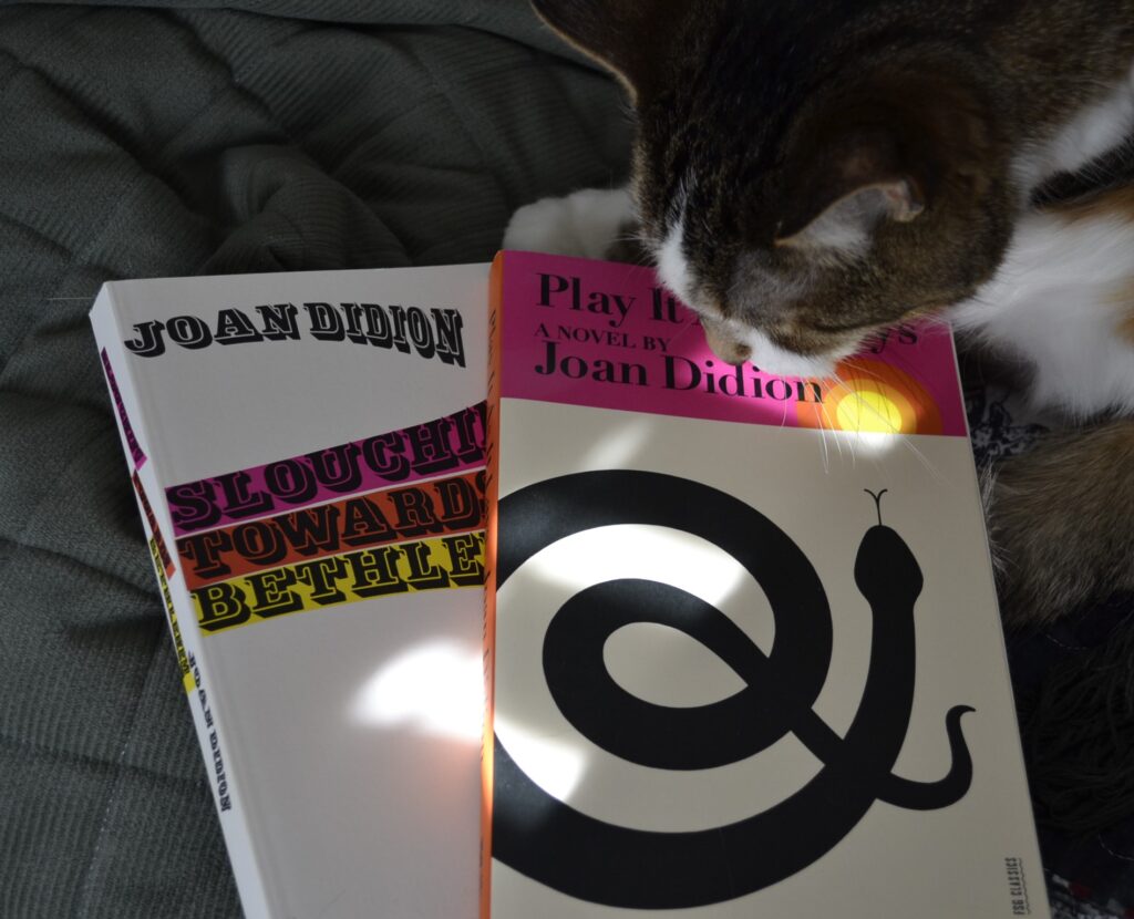 A tabby cat rests her chin on two books: Slouching Towards Bethlehem and Play It As It Lays.