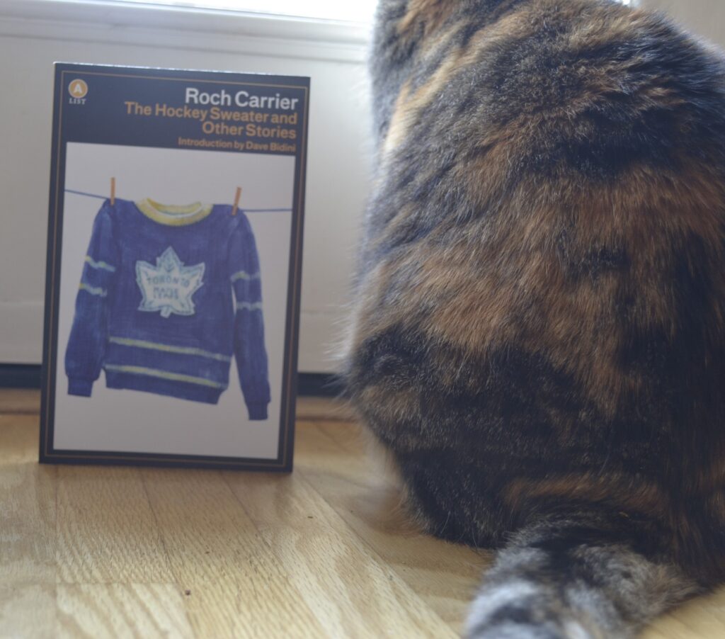 A book sits beside a cat. It is Roch Carrier's The Hockey Sweater.