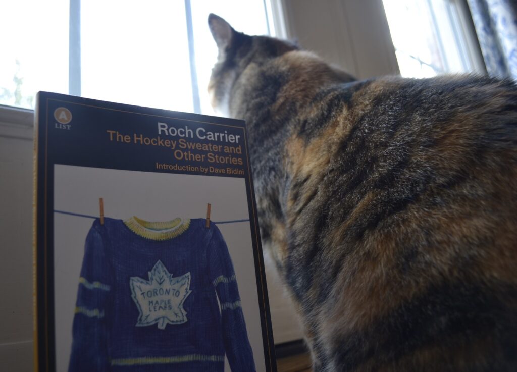 A calico tabby looks out a window. Beside her is Roch Carrier's The Hockey Sweater and Other Stories.