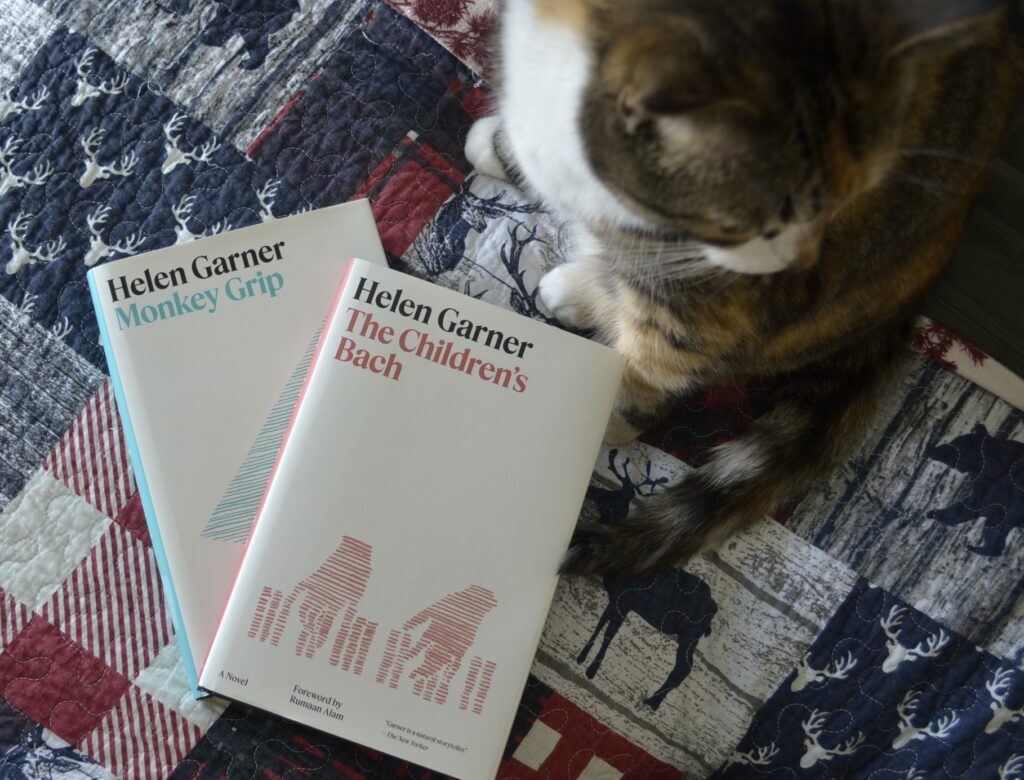Two books by Helen Garner are fanned on a bed with a busy blue quilt. A calico tabby sits beside them.