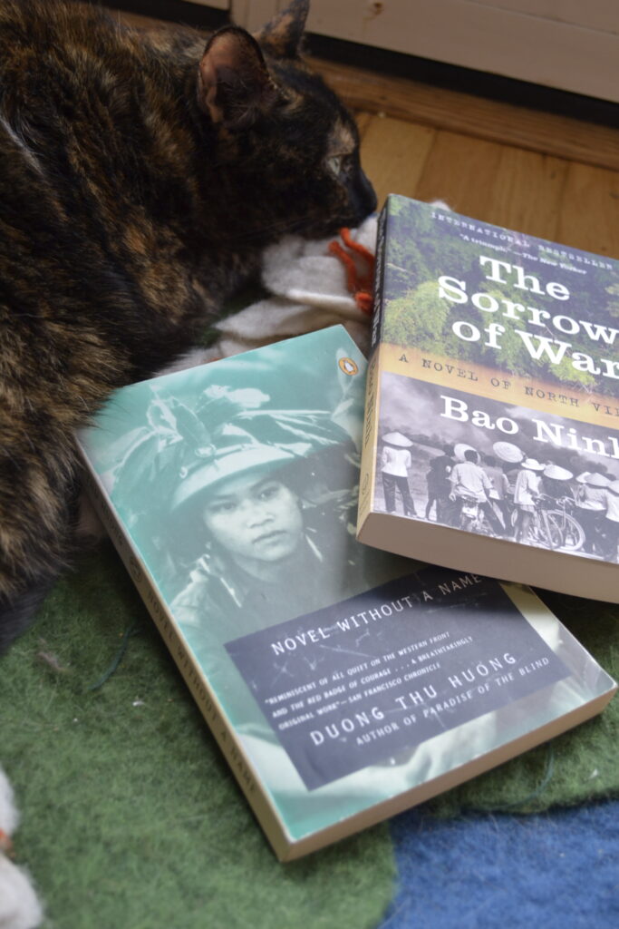A tortoiseshell cat curls around two books: The Sorrow of War and Novel Without a Name.