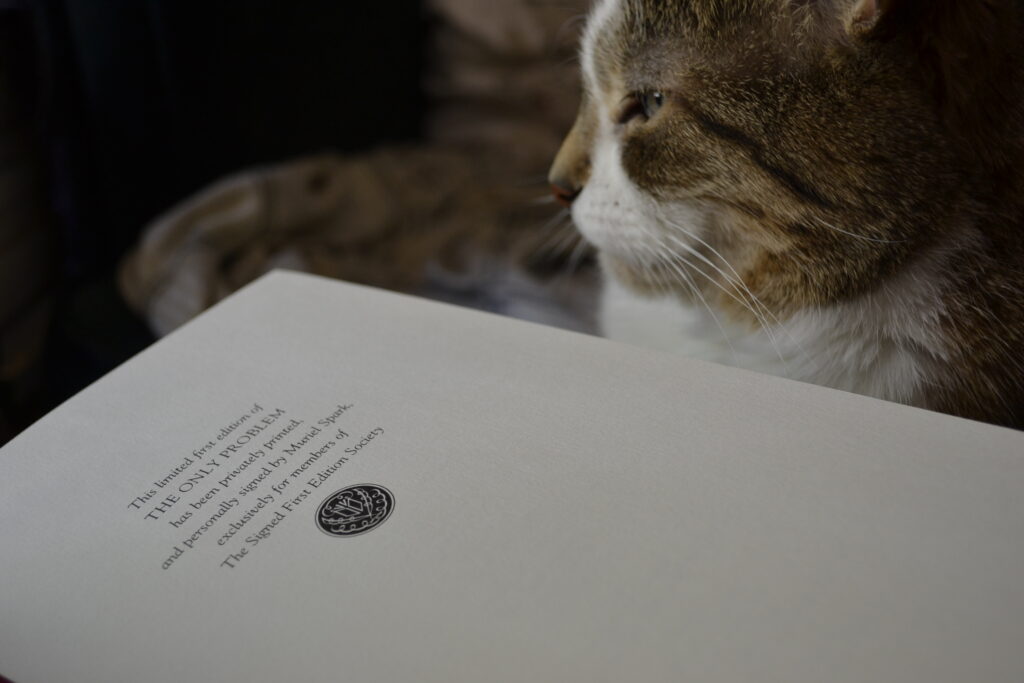 Beside a cat, a book is open. The page says 'This limited first edition of THE ONLY PROBLEM have been privately printed, and personally signed by Muriel Spark, exclusively for members of The Signed First Edition Society.'