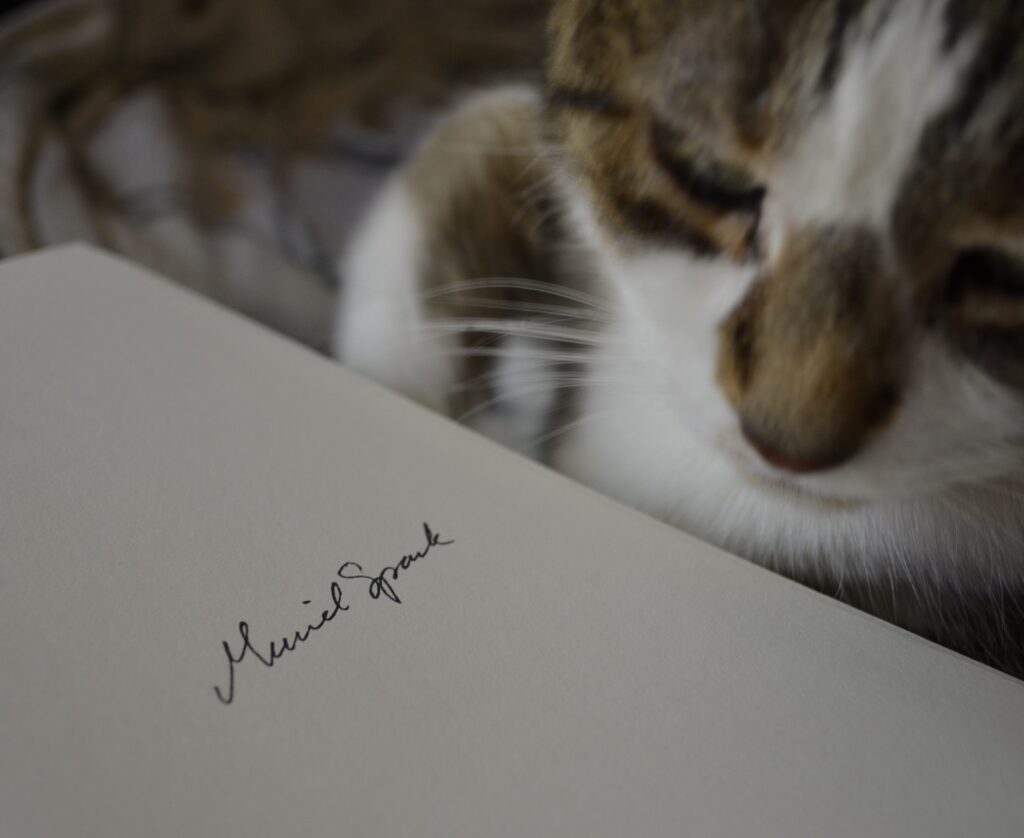 A tabby cat sits beside an open book. On the page is Muriel Spark's signature.