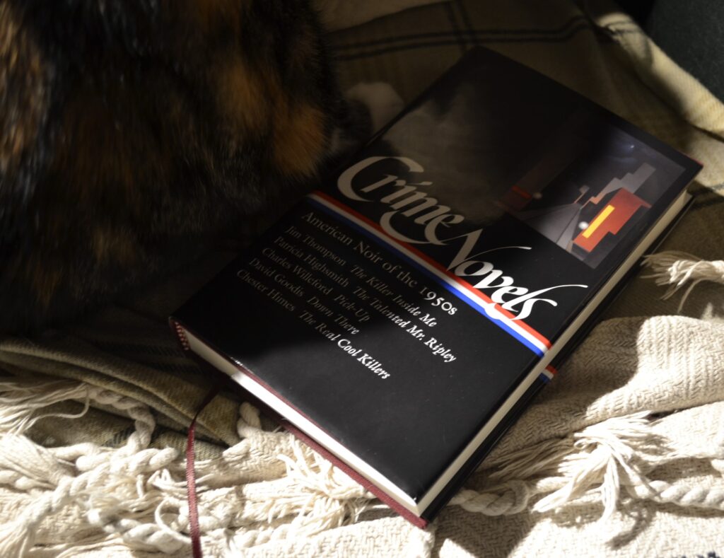 A calico tabby sits on the edge of a book: Crime Novels, American Noir of the 1950s.