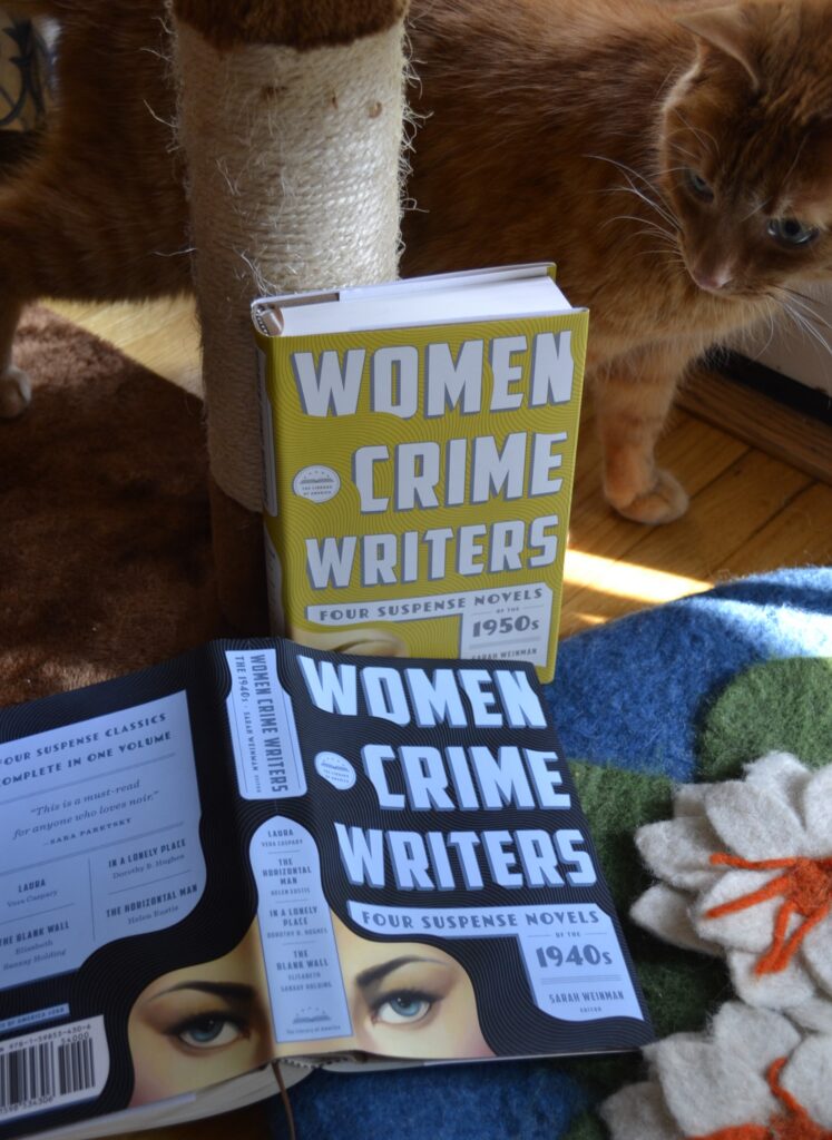 An orange cat curls around a cat tree pole to look at two books: both volumes of Women Crime Writers.