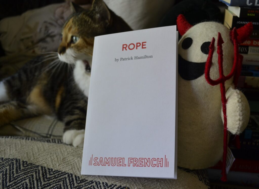 A white ghost toy wearing a read devil costume holds a white book with a red title. A calico tabby yawns behind the book.