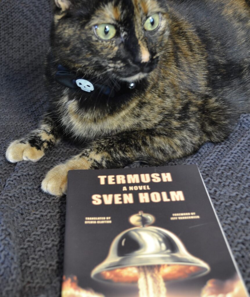 A tortoiseshell cat sits politely wearing a black bow with a skeleton charm in the middle. Below her paws is Termush.