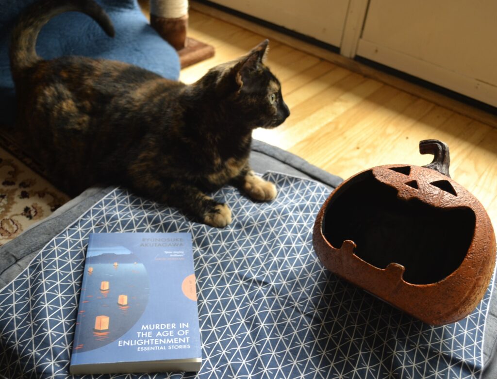 A tortie sits beside a copy of Murder in the Age of Enlightenment by Ryunosuke Akutagawa.