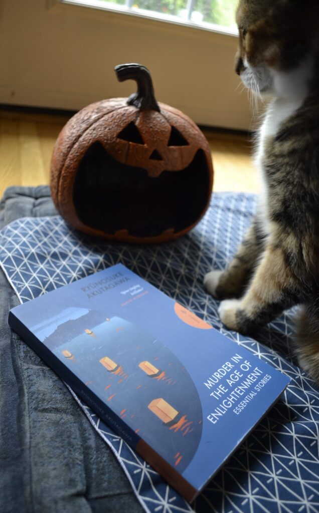 A calico tabby looks down at a grinning jack-o-lantern. Beside her is a blue book: Murder in the Age of Enlightenment by Ryunosuke Akutagawa.