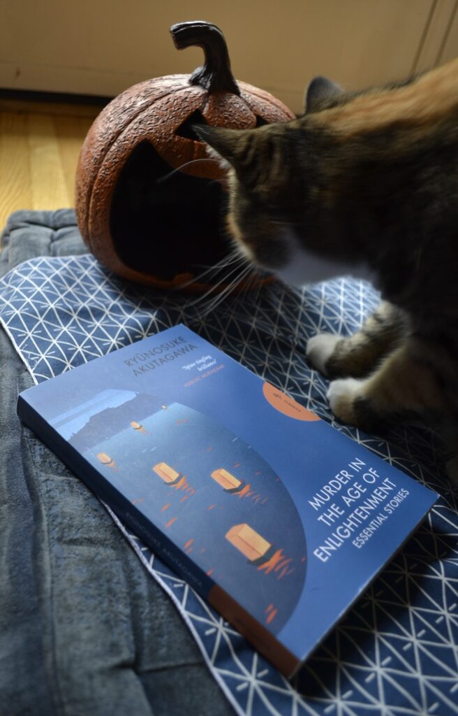 A calico tabby sniffs a blue book with floating lanterns on the cover — Murder in the Age of Enlightenment, Essential Stories by Ryunosuke Akutagawa.