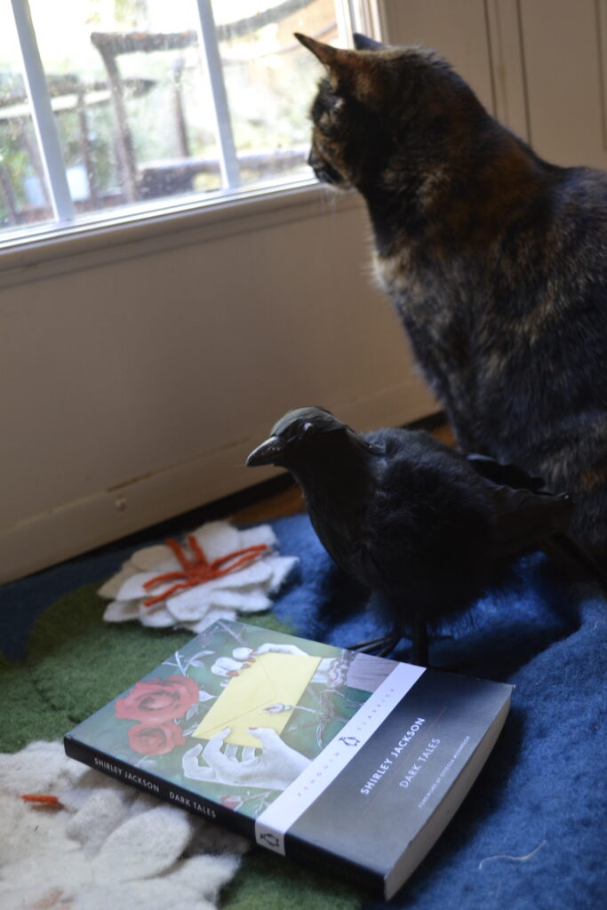 Shirley Jackson's Dark Tales lies on the ground beside a fake crow. A tortie looms in the background.