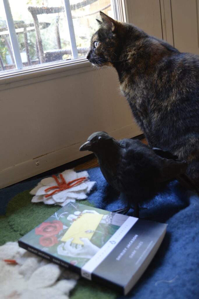 A tortoiseshell cat looks out a window. At her feet are a fake crow and a book.