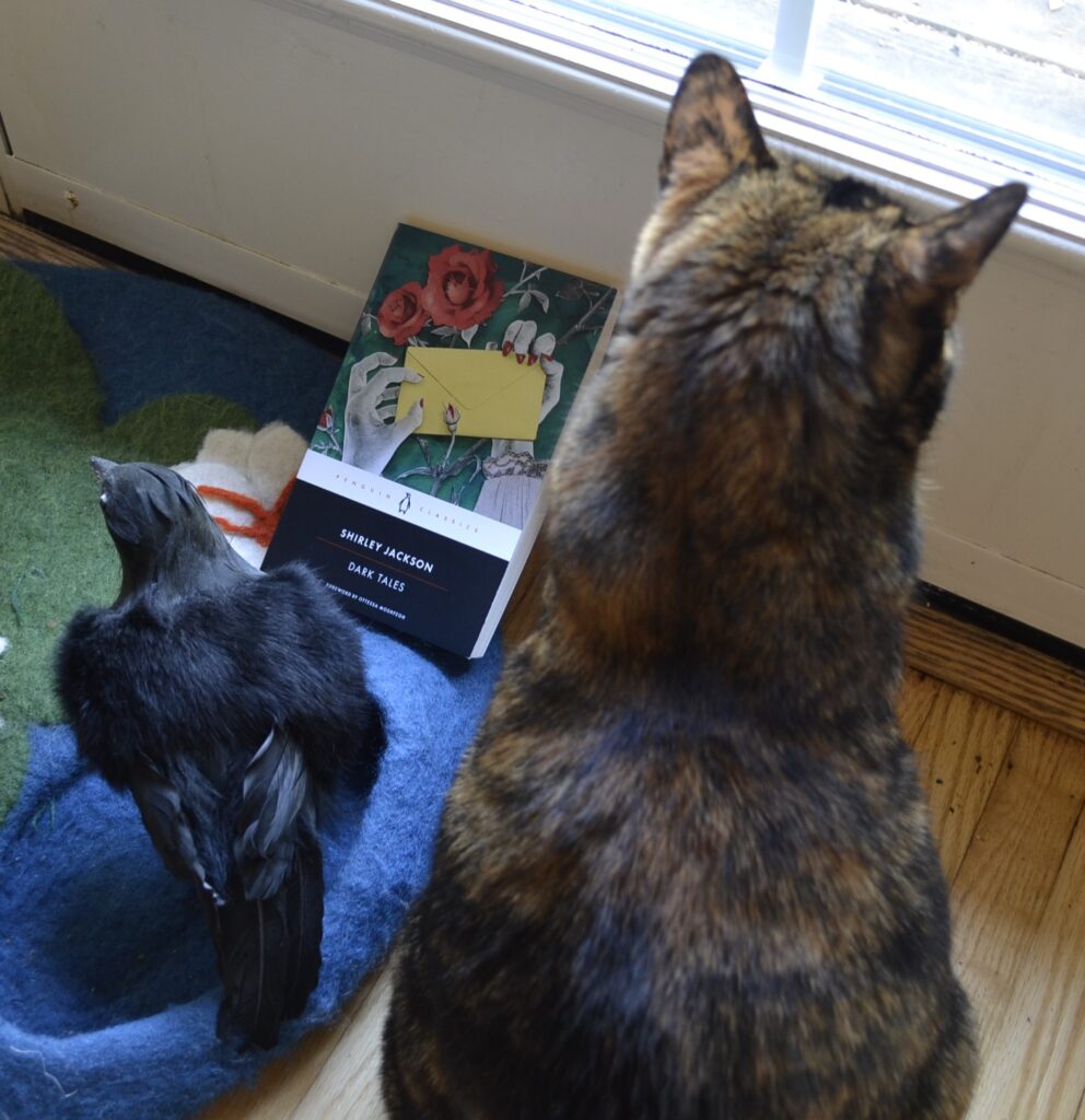 Beside a fake crow and a tortie is a book. The cover features white hands with sharp red nails that are grasping an envelope.