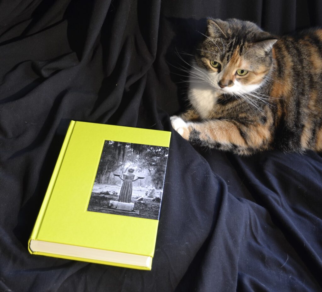 An electric green book has a black-and-white photo on the front: a status of a young woman holding up a plate in each hand. A calico tabby sits behind the book.