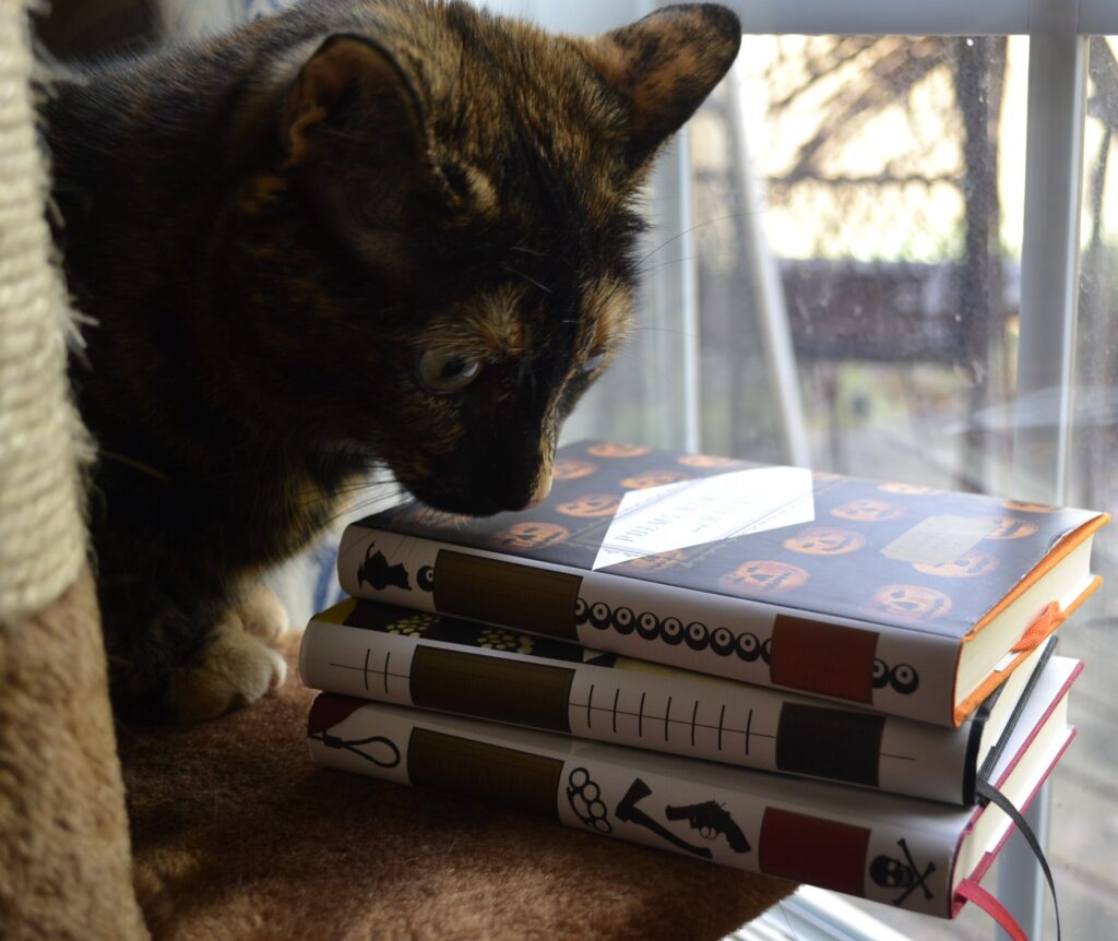 A tortoiseshell cat intensely sniffs a stack of three books with pumpkins, murder weapons, and stitching on the spines.