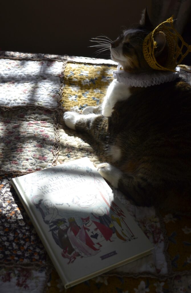 A cat tilts her head back, her tiny crown slipping over one ear. Her ruffled collar has a gold button in the centre front. Alice in Wonderland lies on the bed beside her.