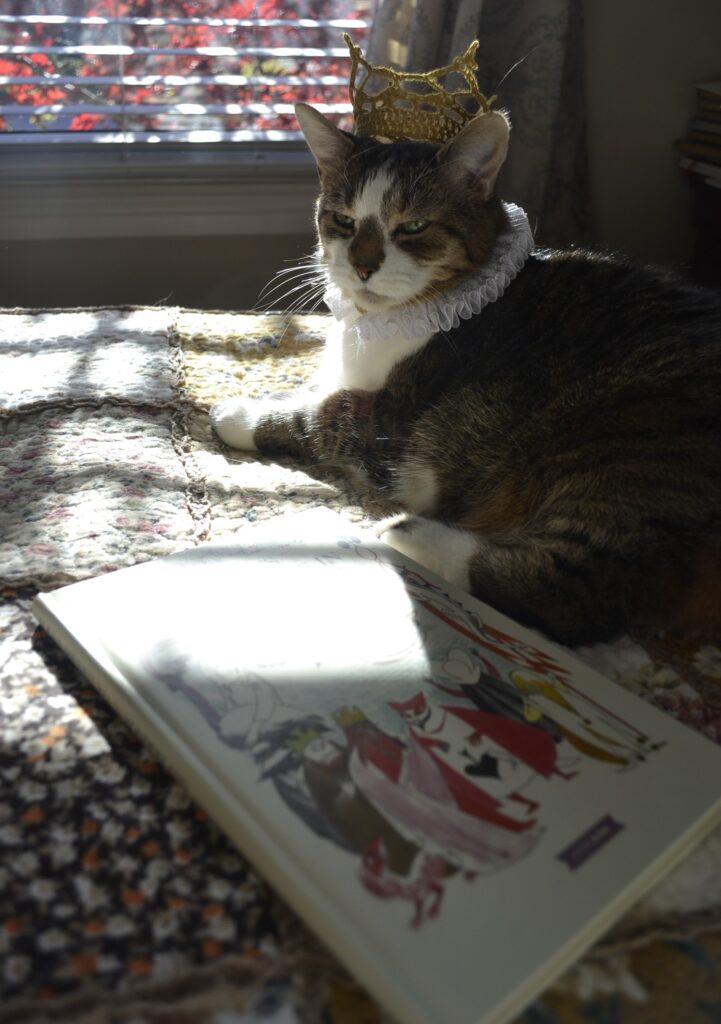 A tabby cat sits majestically on a patchwork blanket. She wears a small crochet crown and a lace Elizabethan-style ruff collar. Beside her is a copy of Alice in Wonderland.