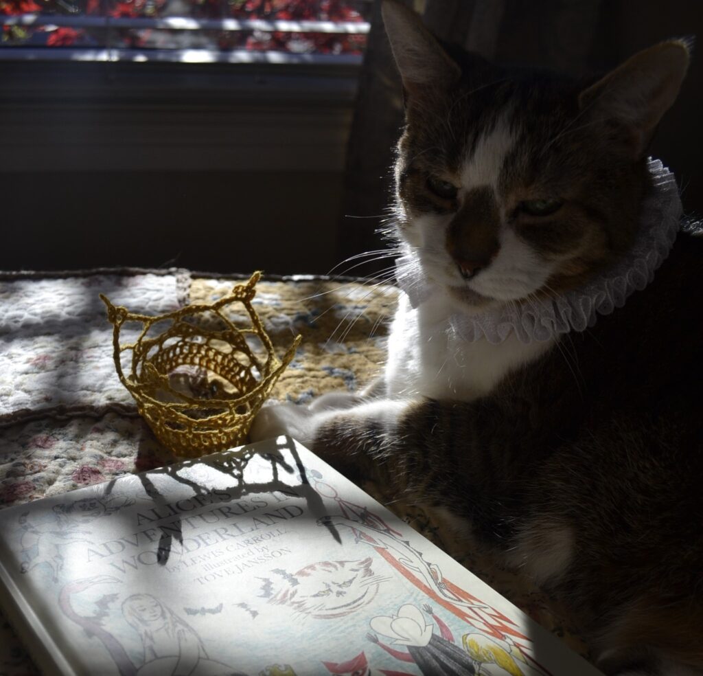 A cat in a ruff sits beside Alice's Adventures in Wonderland. A little crown casts a shadow on the book.