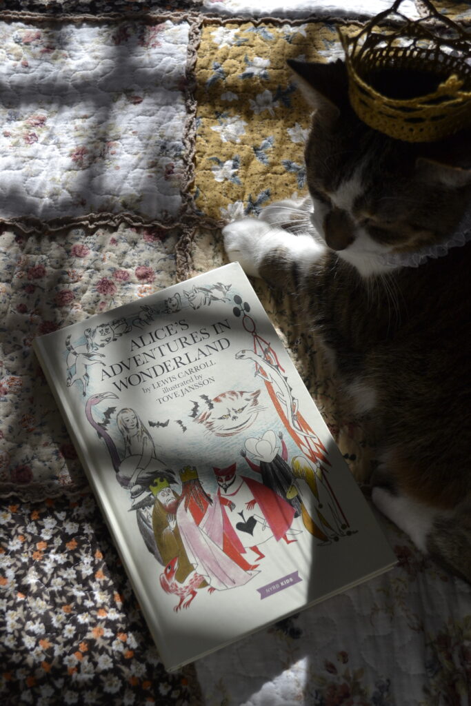 A tabby cat wearing a small crochet crown curls around a copy of Alice's Adventures in Wonderland, illustrated by Tove Jansson.